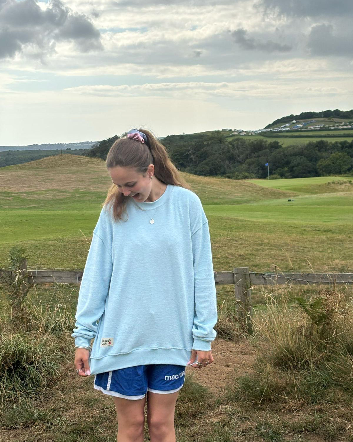 Person wearing a light blue sweatshirt and blue shorts standing in a grassy field with hills and houses in the background.