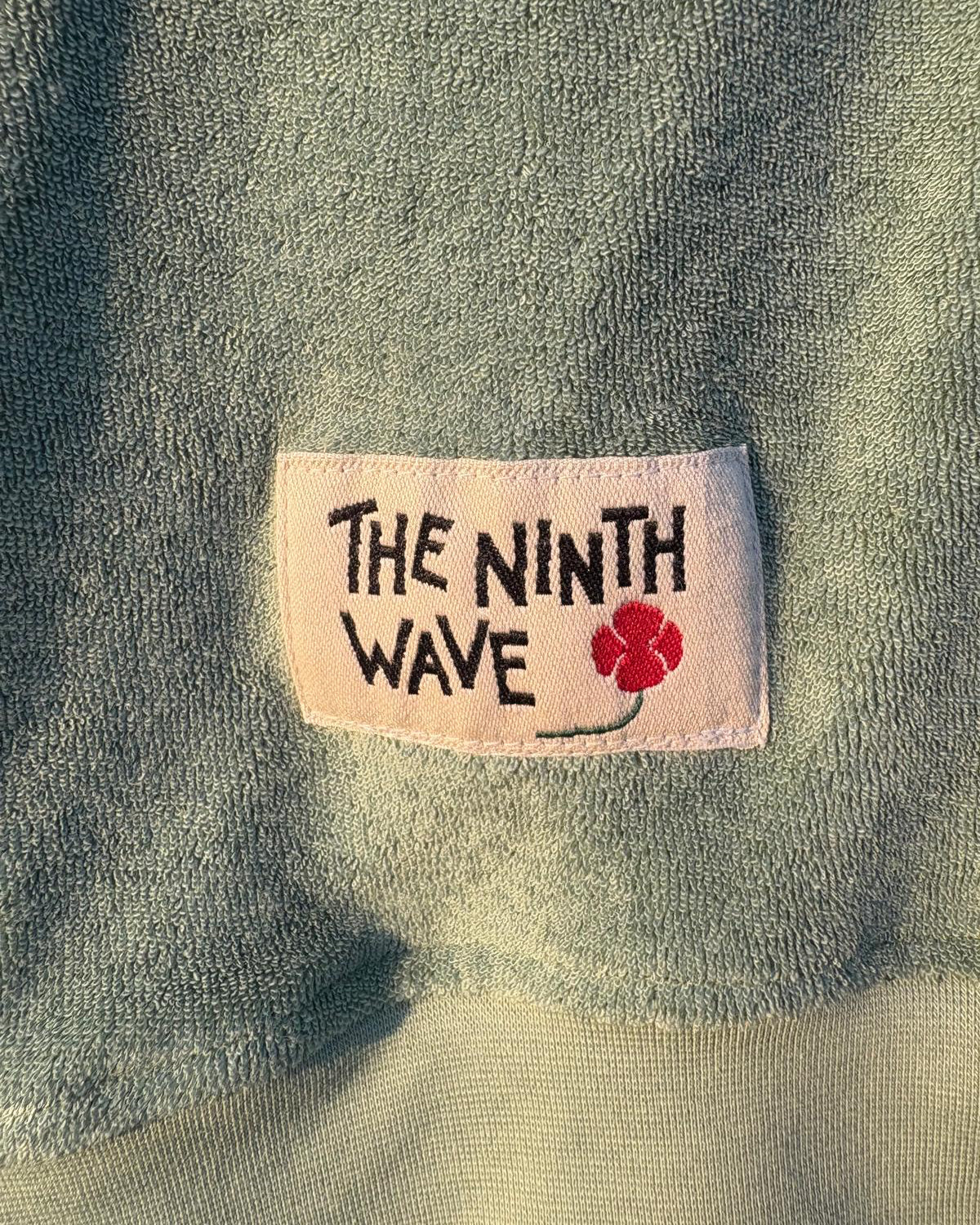 Close-up of a Baby Blue Towelling Jumper with 'The Ninth Wave' label featuring a flower design.
