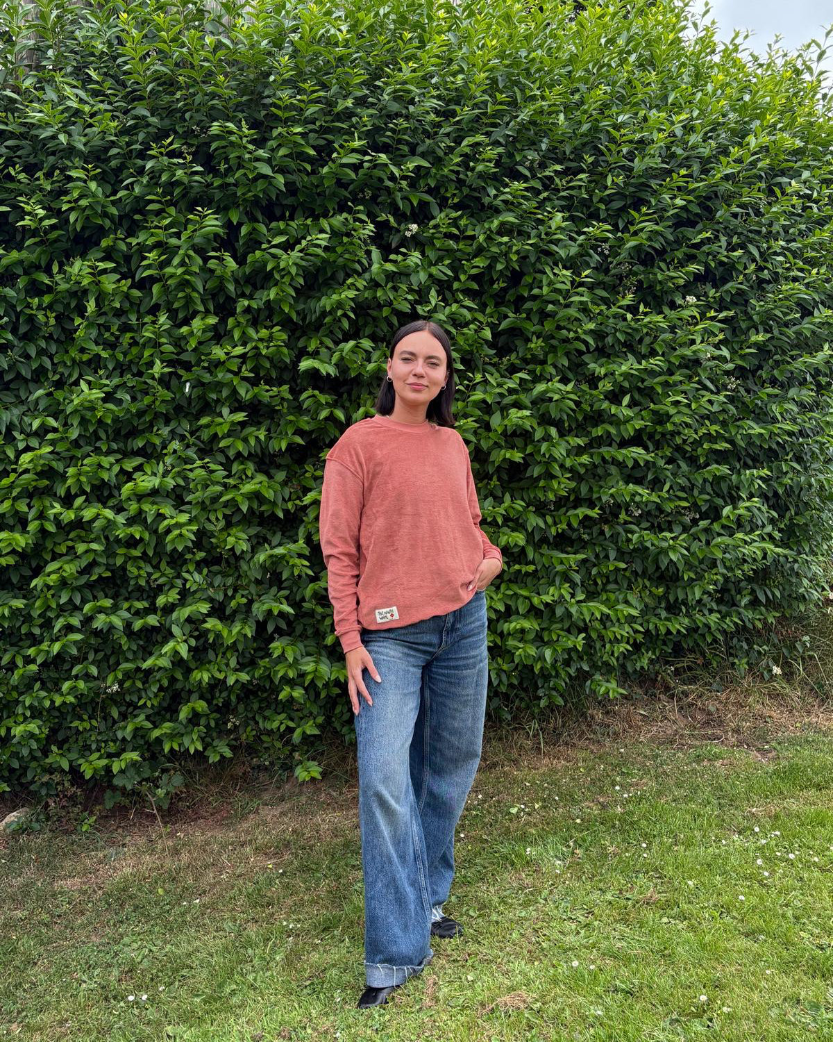 Person wearing a faded terracotta sweater and blue jeans standing in front of a green hedge.