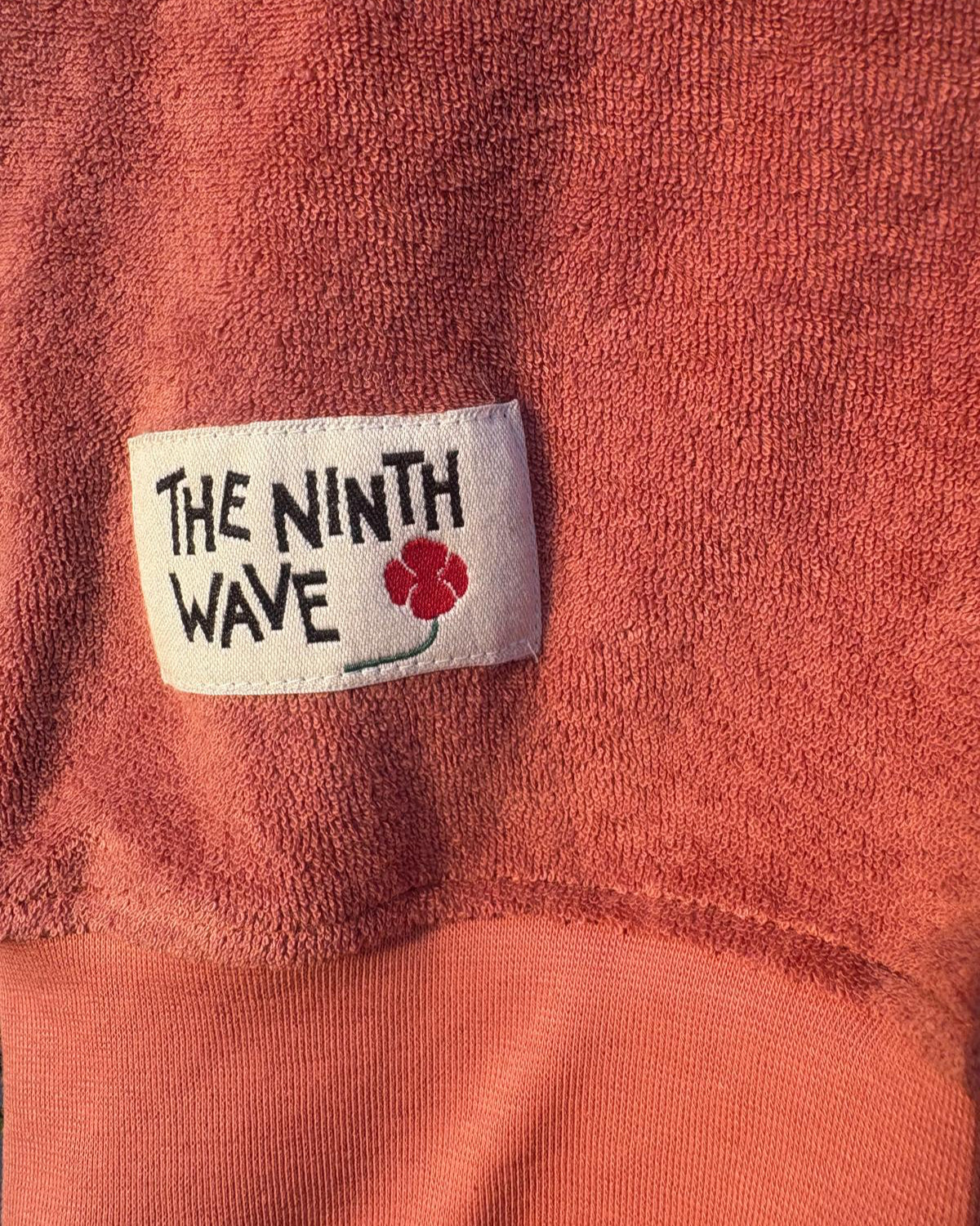 Close-up of a Faded Terracotta Towelling Jumper with 'The Ninth Wave' label featuring a flower design.