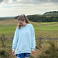 Person wearing a light blue sweatshirt and blue shorts standing in a grassy field with hills and houses in the background.