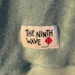 Close-up of a Baby Blue Towelling Jumper with 'The Ninth Wave' label featuring a flower design.