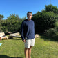 Man standing in a garden wearing a dark navy jumper with greenery and a clear blue sky behind him.