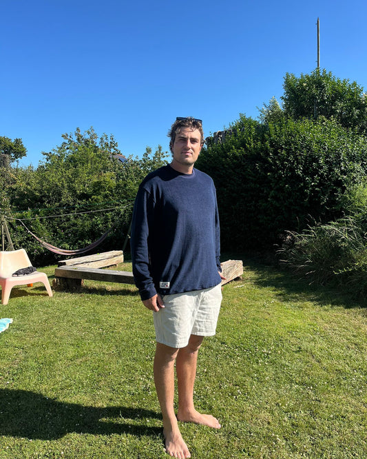 Man standing in a garden wearing a dark navy jumper with greenery and a clear blue sky behind him.