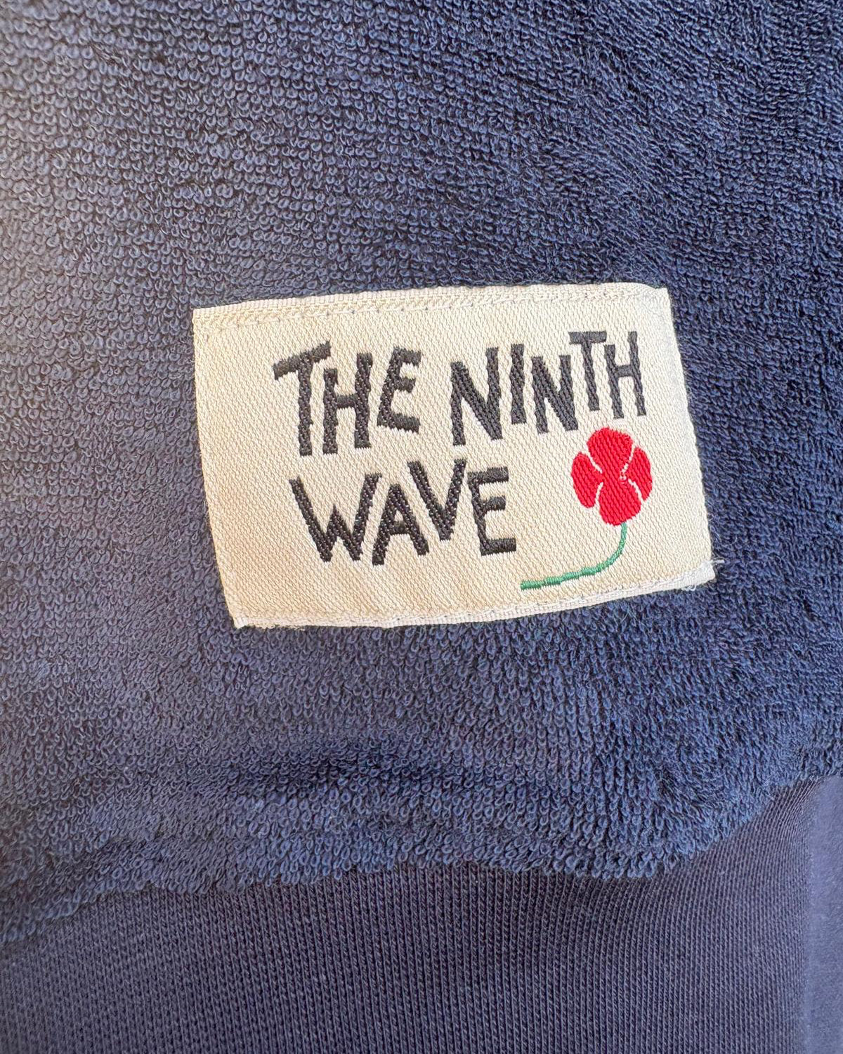 Close-up of a Dark Navy Towelling Jumper with 'The Ninth Wave' label featuring a flower design.