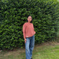 Person wearing a faded terracotta sweater and blue jeans standing in front of a green hedge.