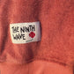 Close-up of a Faded Terracotta Towelling Jumper with 'The Ninth Wave' label featuring a flower design.