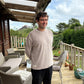 Man standing on a wooden deck with outdoor furniture and greenery in the background. He's wearing a mushroom coloured jumper.