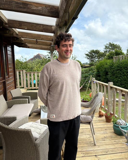 Man standing on a wooden deck with outdoor furniture and greenery in the background. He's wearing a mushroom coloured jumper.